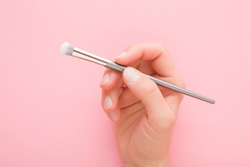 Young adult woman hand fingers holding and showing new makeup brush with soft bristles on light pink table background. Pastel color. Female beauty product. Closeup. Top down view.