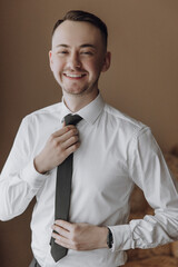 A man is wearing a black tie and smiling. He is wearing a white shirt and is holding a tie in his hands