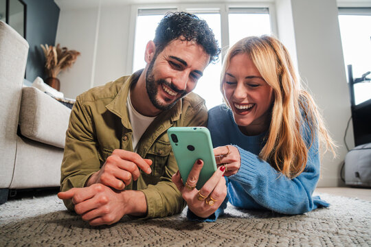 Adult couple watching funny videos together on a cellphone social media app lying on the floor at home. Young caucasian wife and happy husband on the carpet browsing on internet using a smart phone