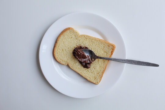 One slice of white bread and one tablespoon of chocolate spread, on white plate, isolated on white background, flat lay or top view