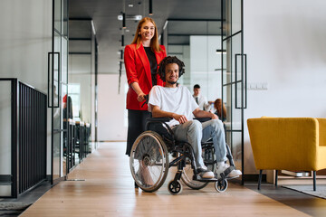 A business leader with her colleague, an African-American businessman who is a disabled person, pass by their colleagues who work in modern offices