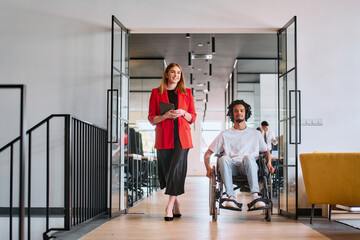 A business leader with her colleague, an African-American businessman who is a disabled person, pass by their colleagues who work in modern offices