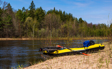 On a clear spring day, a yellow packraft with an oar, loaded with a bicycle and equipment, lies on the sandy bank, ready for rafting down a forest river.