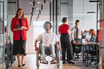A business leader with her colleague, an African-American businessman who is a disabled person, pass by their colleagues who work in modern offices