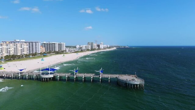 Scenic view of Pompano Beach showcasing beautiful coastline and pier extending into ocean, nearby buildings under clear blue sky. Concept of recreation and landscape