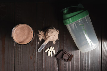 Protein milkshake cocktail in a glass, plastic measuring spoon with whey protein powder, white pills or capsules of amino acids, chocolate cubes on a dark wooden background, top view