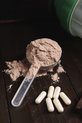 Plastic measuring spoon with whey protein powder, milkshake cocktail in a glass, blended protein drink, white pills or capsules, chocolate cubes on a dark wooden background