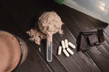 Plastic measuring spoon with whey protein powder, milkshake cocktail in a glass, blended protein drink, white pills or capsules, chocolate cubes on a dark wooden background, top view