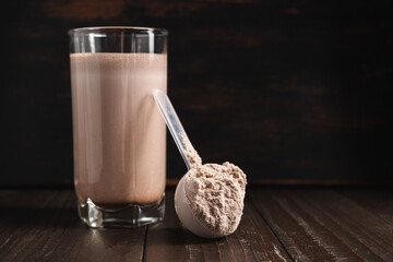 Plastic measuring spoon with whey protein powder, chocolate milkshake cocktail in a glass, blended...