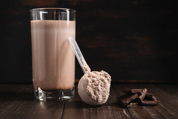 Plastic measuring spoon with whey protein powder, milkshake cocktail in a glass, blended protein drink and chocolate cubes on a dark wooden background