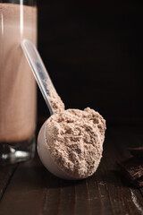 Plastic measuring spoon with whey protein powder, chocolate milkshake cocktail in a glass, blended protein drink on a dark wooden background