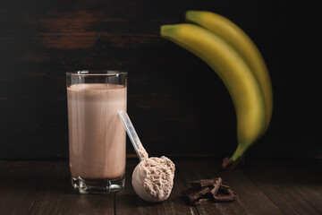 Plastic measuring spoon with whey protein powder, milkshake cocktail in a glass, blended protein drink, chocolate cubes and banana fruit on a dark wooden background