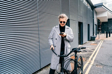 Stylish male in coat and sunglasses using mobile phone while walking with retro bicycle in city....