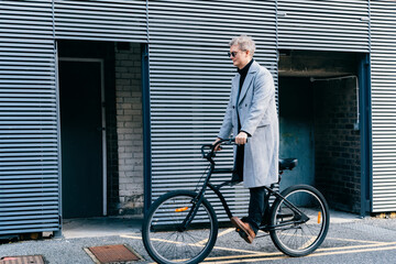 Stylish male in checked coat and sunglasses riding on retro bicycle near gray urban wall in sunny...