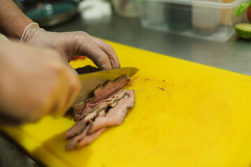 Kitchen worker expertly cuts roasted pork with sharp knife on cutting board. Culinarian crafts thin...
