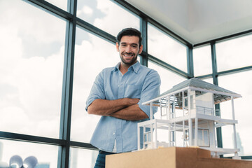 Portrait of architect engineer in casual outfit smile at camera while crossing arms. Businessman looking at camera and standing with arms folded near house model, architectural model. Tracery.