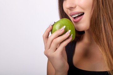 A young woman smiles as she eats a green apple. She has braces on her teeth, long brown hair, and...