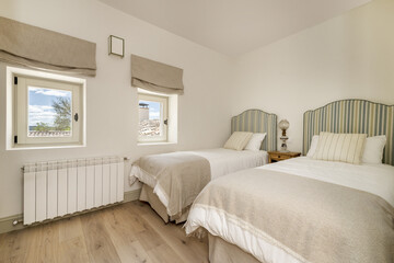 A bedroom with two single beds with headboards upholstered in striped fabric, matching cushions and pillows, and two square windows with views