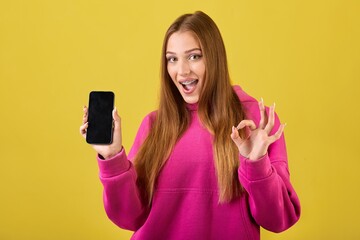 Expressive female with braces on teeth approving something and holding mobile phone in hand. Excited young woman in casual clothes showing okay sign and looking at camera.