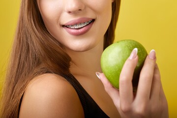 A young woman with braces on her teeth is holding a green apple in front of her mouth. She has long...