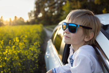 7-8 year old boy in blue sunglasses looks out the open car window and examines the nature around...