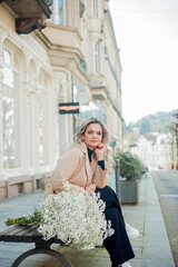 Portrait of beautiful young blonde woman with bouquet of white flowers in the center of European city. Happy model in casual spring clothes. Holiday.