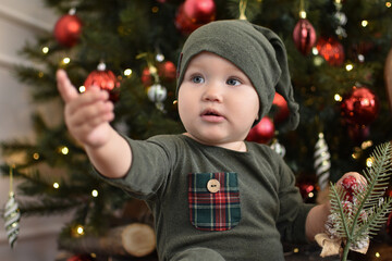 Baby 1 year old sitting in chair with Christmas tree and lights on background in room. Merry Christmas. Holiday season