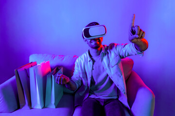 Caucasian man with vr glass posing pointing finger while sitting at sofa with shopping bags. Person...