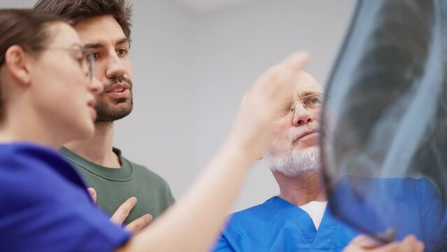 A couple of confident doctors a girl and an elderly man with a gray beard and glasses reassure a guy showing good results of fluorography on an image in a modern clinic