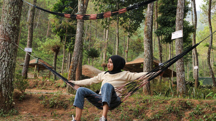Pensive smiling young woman with headscarf resting in comfortable hammock at green garden.