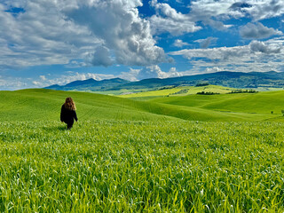 Obraz premium A woman walks through lush green fields under a clear blue sky, Tuscany hills, Italy.