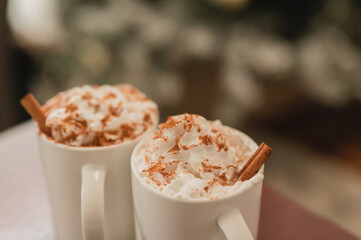 Close up of hot chocolate in a white ceramic mug with whipped cream and cinnamon powder & cinnamon sticks against a Christmas tree scene