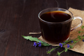 close-up of hot chicory drink in glass cup with blue chicory flowers with sackcloth on dark brown wooden background. healthy. coffee substitute. copy space.