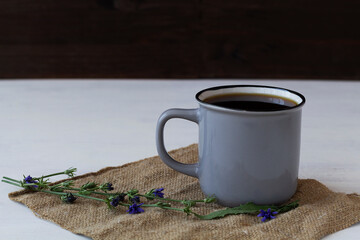 Herbal beverage, coffee substitute. Grey Cup of delicious chicory drink and blue chicory flowers on white table with sackcloth. hot and healthy drinks. copy space
