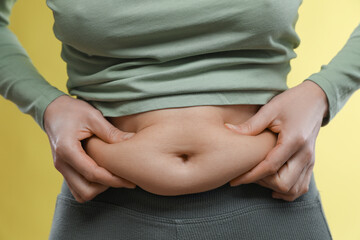 Woman touching belly fat on yellow background, closeup. Overweight problem
