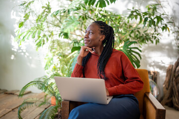 Dreaming African American woman distracted from work on laptop looks at window sits on chair, home...