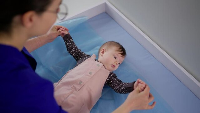 From above over the shoulder a confident brunette girl doctor in glasses finds a common language and checks a little baby girl in a pink overalls for the correct development in a modern clinic at a