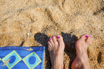 A woman's feet are on the beach with a towel and a bottle of nail polish