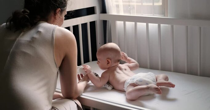 A view from the back of a mother who is sitting by a crib in which lies a infant baby in a diaper and with a bunny toy. The mother gently touches the child and wants to put him to sleep. Mothers Day.