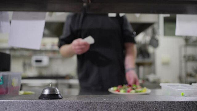 A professional male chef in a black uniform places a ready-made salad order on the serving table and rings the bell to notify the waiters
