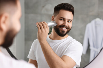 Handsome young man trimming beard near mirror in bathroom