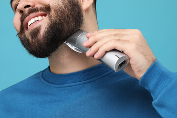 Handsome young man trimming beard on light blue background, closeup