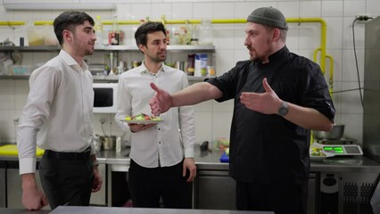 A confident male chef in a black uniform tells two young guys waiters how to properly take out dishes and work in the kitchen with staff in a restaurant