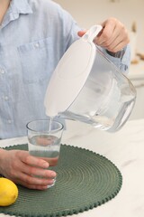Woman pouring water from filter jug into glass at white marble table in kitchen, closeup