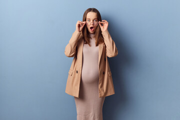 Shocked pregnant woman in beautiful dress and jacket posing isolated over blue background taking...