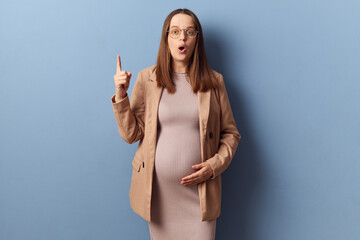 Inspired surprised pregnant woman in beautiful dress and jacket posing isolated over blue...