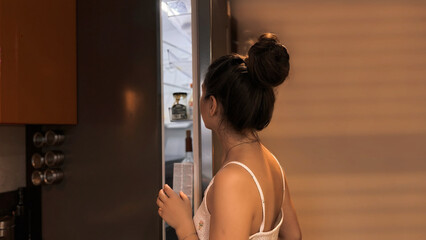 A girl with dark hair tied up in a bun and wearing pajamas looks into the refrigerator. A hungry young woman opened the refrigerator and chooses food, eats at home