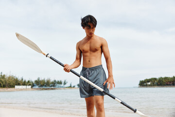 Naklejka premium Active Asian Man Paddleboarding: Summer Fun and Adventure on Beach with Sunset