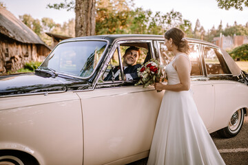 Valmiera, Latvia - August 19, 2023 - A bride stands beside a vintage car, handing a bouquet to the groom who smiles from inside the car.