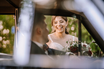 Valmiera, Latvia - August 19, 2023 - Bride smiling at groom in vintage car, lace dress, happy...